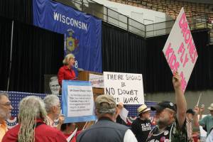 Tammy Baldwin speaking at Fighting Bob Fest, 2013, while peace activists hold signs and protest her unwillingness to say she'll vote no on any war or action against Syria.  Photo by Callen Harty.